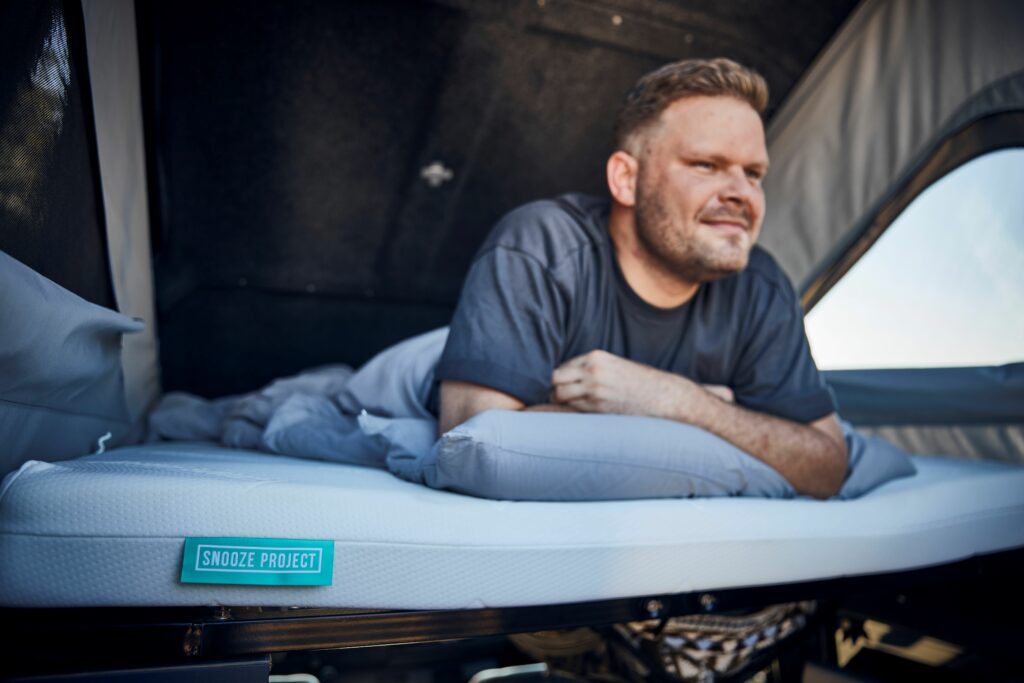 Ein Mann liegt auf dem Bauch auf einem Bett in einem Zelt oder Wohnwagen und ruht sich auf einer grauen Dachzeltmatratze mit der Aufschrift Snooze Project aus. Er lächelt und blickt in das helle Tageslicht hinaus.