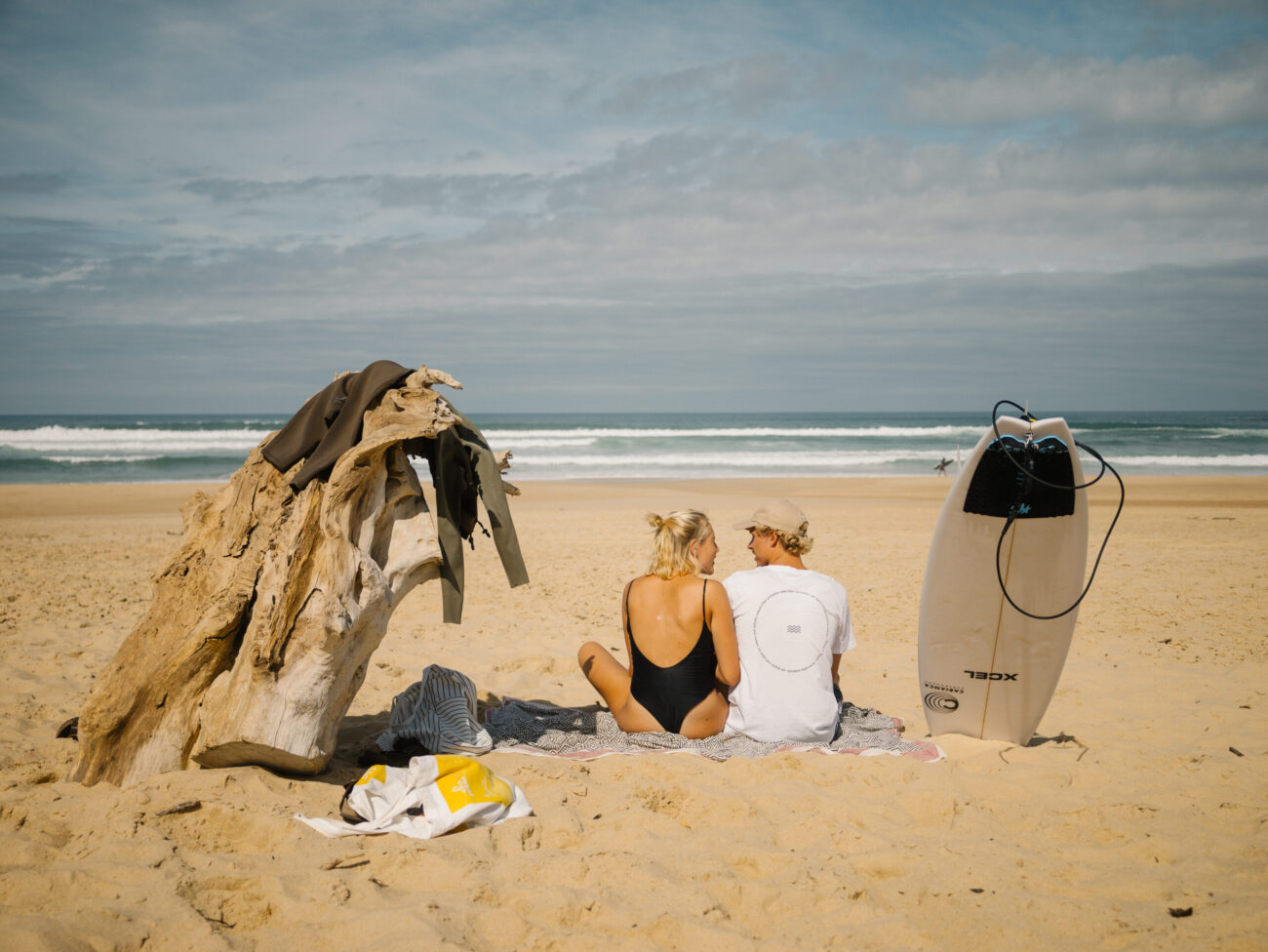 Ein Pärchen sitzt auf einem Strandtuch mit Blick auf den Ozean, Surfbrett und Treibholz in der Nähe, und träumt von ihrem nächsten Abenteuer - vielleicht ein Bus umbauen, um neue Küsten zu erkunden und besser zu schlafen unter dem teilweise bewölkten Himmel.