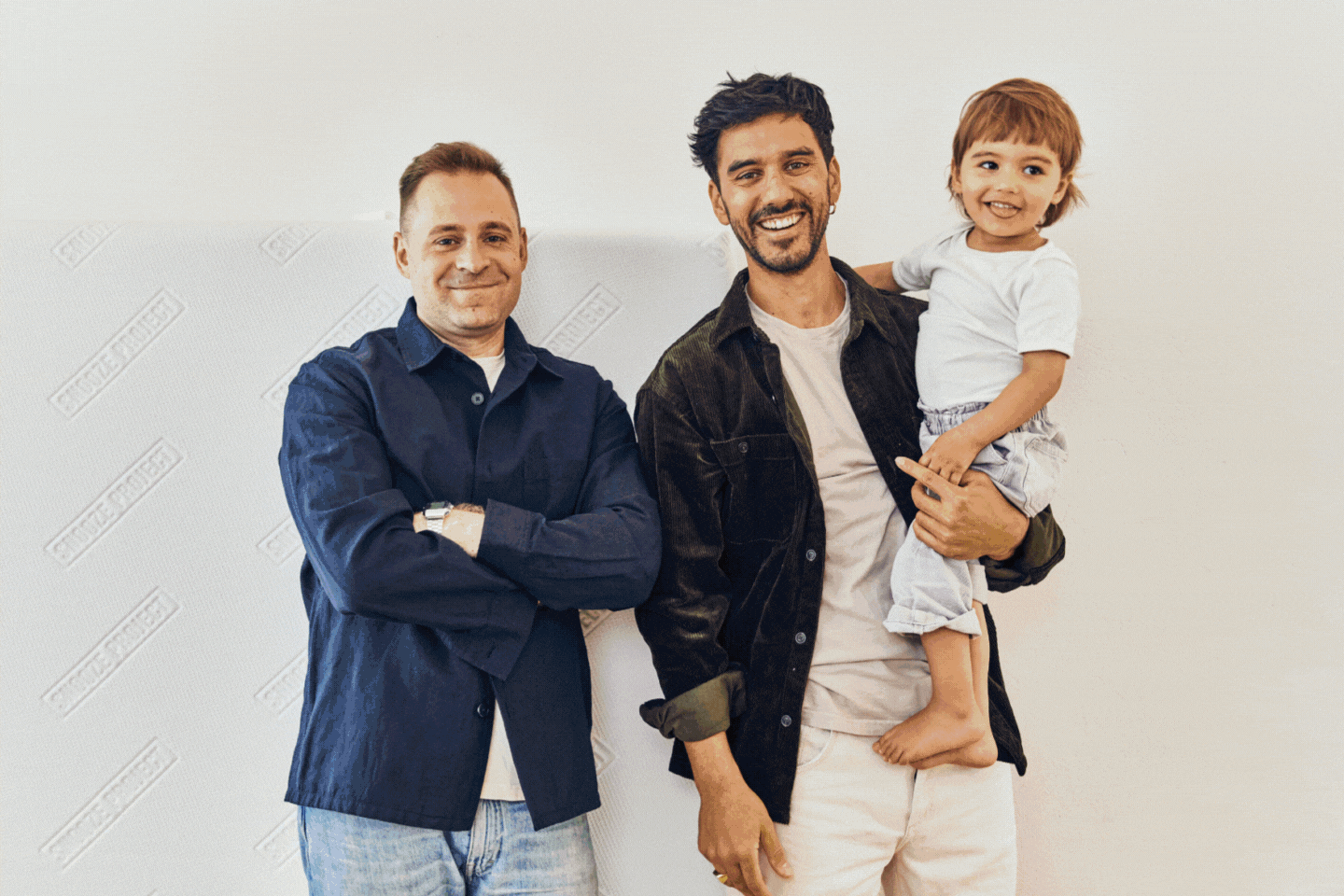 Zwei Männer stehen zusammen und lächeln in die Kamera; einer hält ein kleines Kind, das ebenfalls lächelt. Die Gruppe ist leger gekleidet und posiert vor einem schlichten, hellen Hintergrund.