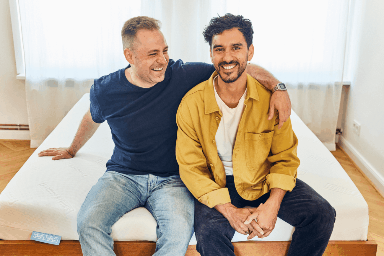 Zwei Männer sitzen auf einem Bett und lächeln. Der eine trägt ein marineblaues T-Shirt und Jeans und hat seinen Arm um den anderen gelegt, der ein gelbes Hemd und eine schwarze Hose trägt. Sie wirken glücklich und entspannt in einem hellen Raum mit weißen Vorhängen.