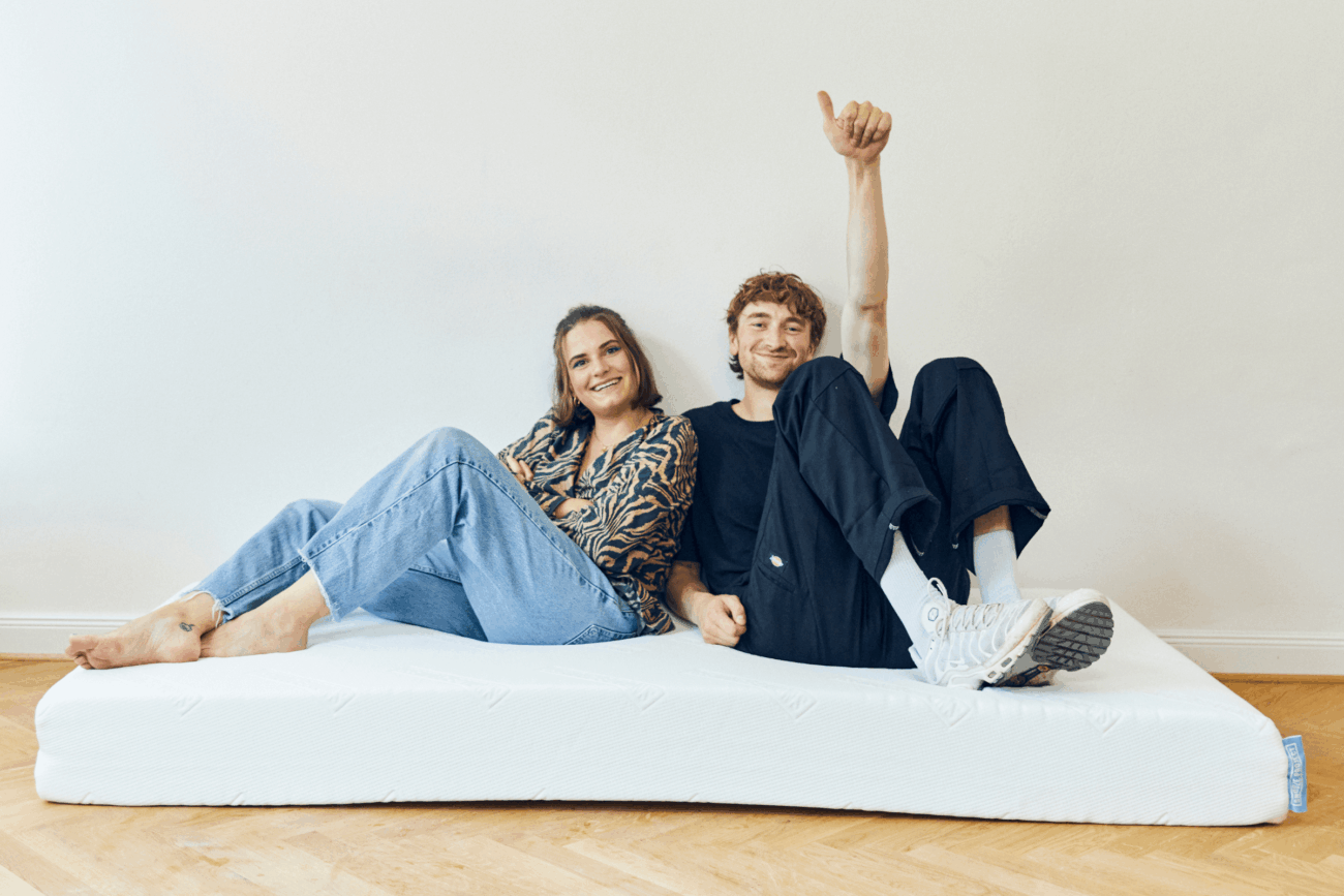 Eine Frau und ein Mann sitzen lässig auf einer weißen Matratze auf einem Holzboden und lächeln. Der Mann zeigt den Daumen nach oben, und beide wirken entspannt und glücklich vor dem Hintergrund einer schlichten weißen Wand.