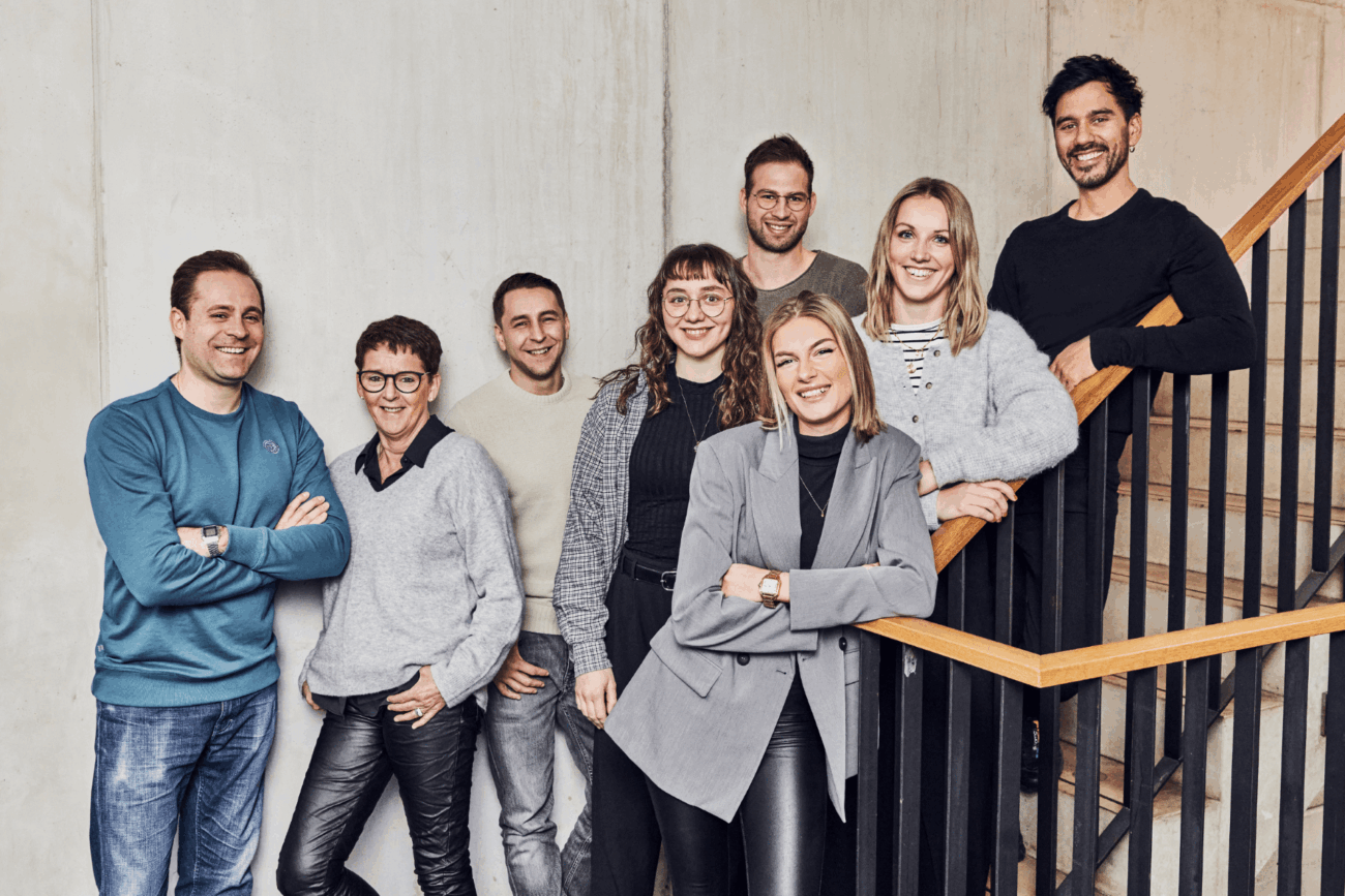 Acht Personen posieren und lächeln gemeinsam in einem Innenraum an einer Treppe, in Freizeit- und Business-Casual-Kleidung, mit einer hellen Wand im Hintergrund.