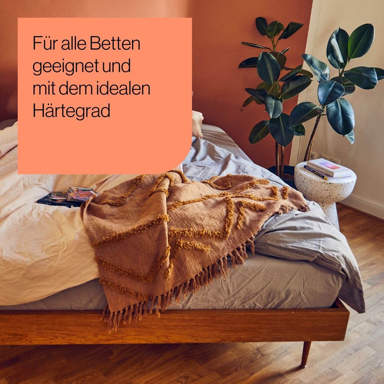 24 Ein ordentlich gemachtes Holzbett mit grauer und beiger Bettwäsche und einer braunen Überwurfdecke steht neben einem weißen Beistelltisch mit einer Pflanze. Der deutsche Text darüber lautet: Für alle Betten geeignet und mit dem idealen Härtegrad.