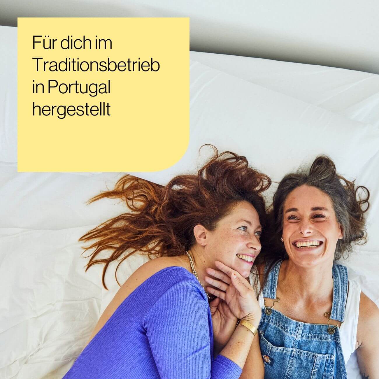 4 Zwei Frauen liegen auf einem Bett, lächeln und schauen sich an. Die eine trägt ein blaues Oberteil, die andere einen Jeans-Overall. In einem gelben Kasten in der Ecke steht ein deutscher Text: Für dich im Traditionsbetrieb in Portugal hergestellt.