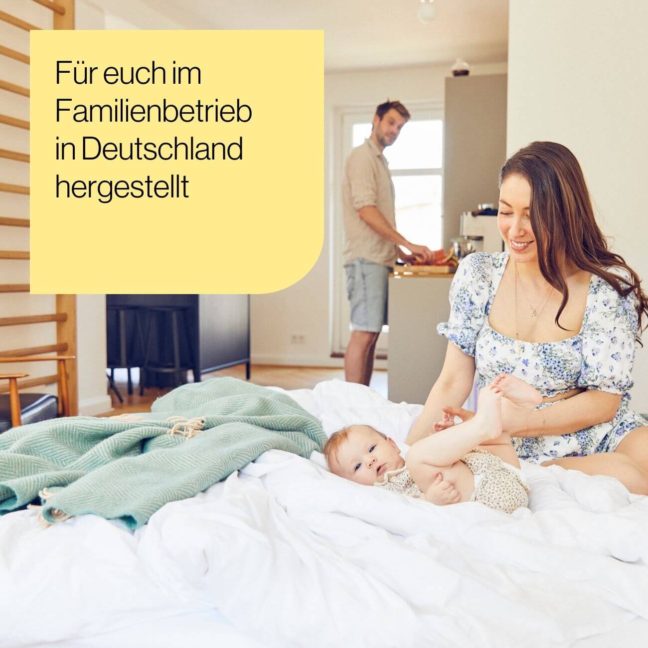 5 Eine lächelnde Frau spielt mit einem Baby auf einem Bett in einem hellen Raum, während ein Mann im Hintergrund in der Küche steht. Der deutsche Text auf dem gelben Kasten lautet: Für euch im Familienbetrieb in Deutschland hergestellt.