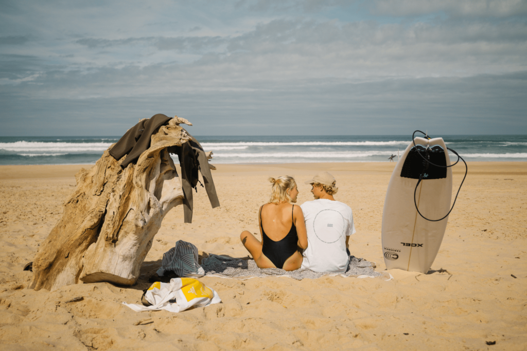 Ein Paar sitzt auf einer Decke und einem Kissen an einem Sandstrand mit Blick auf den Ozean. Ein Surfbrett und Treibholz mit Kleidung hängen in der Nähe. Die Szene im Freien zeigt einen teilweise bewölkten Himmel und im Hintergrund rollende Wellen.