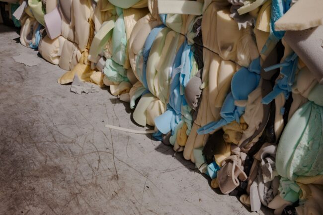 Große, komprimierte Bündel verschiedenfarbiger Schaumstoffstücke, darunter blaue, gelbe, cremefarbene und grüne, gestapelt auf einem Betonboden mit sichtbaren Abnutzungsspuren und Kratzern.