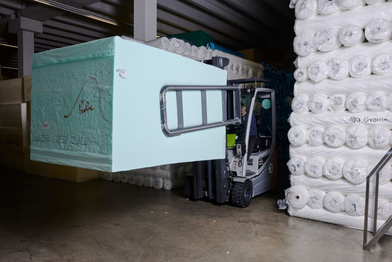 Ein Gabelstapler transportiert einen großen rechteckigen Schaumstoffblock durch ein Lager, das mit gestapelten Rollen aus weißem Material in Regalen und auf dem Boden ausgekleidet ist.