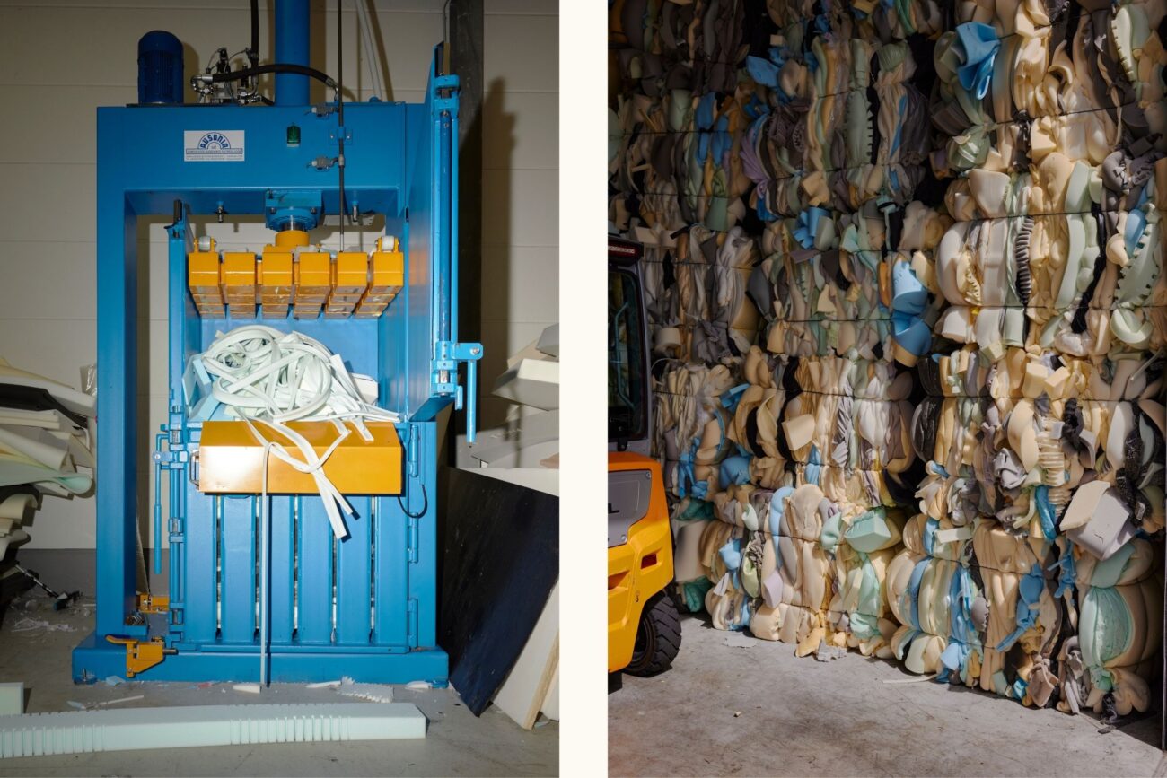 Eine blaue industrielle Schaumstoffpresse verdichtet links Schaumstoffreste; rechts sind zahlreiche große Ballen mit gepressten Schaumstoffstücken entlang einer Wand gestapelt, wobei in der Ecke ein gelber Gabelstapler teilweise sichtbar ist.