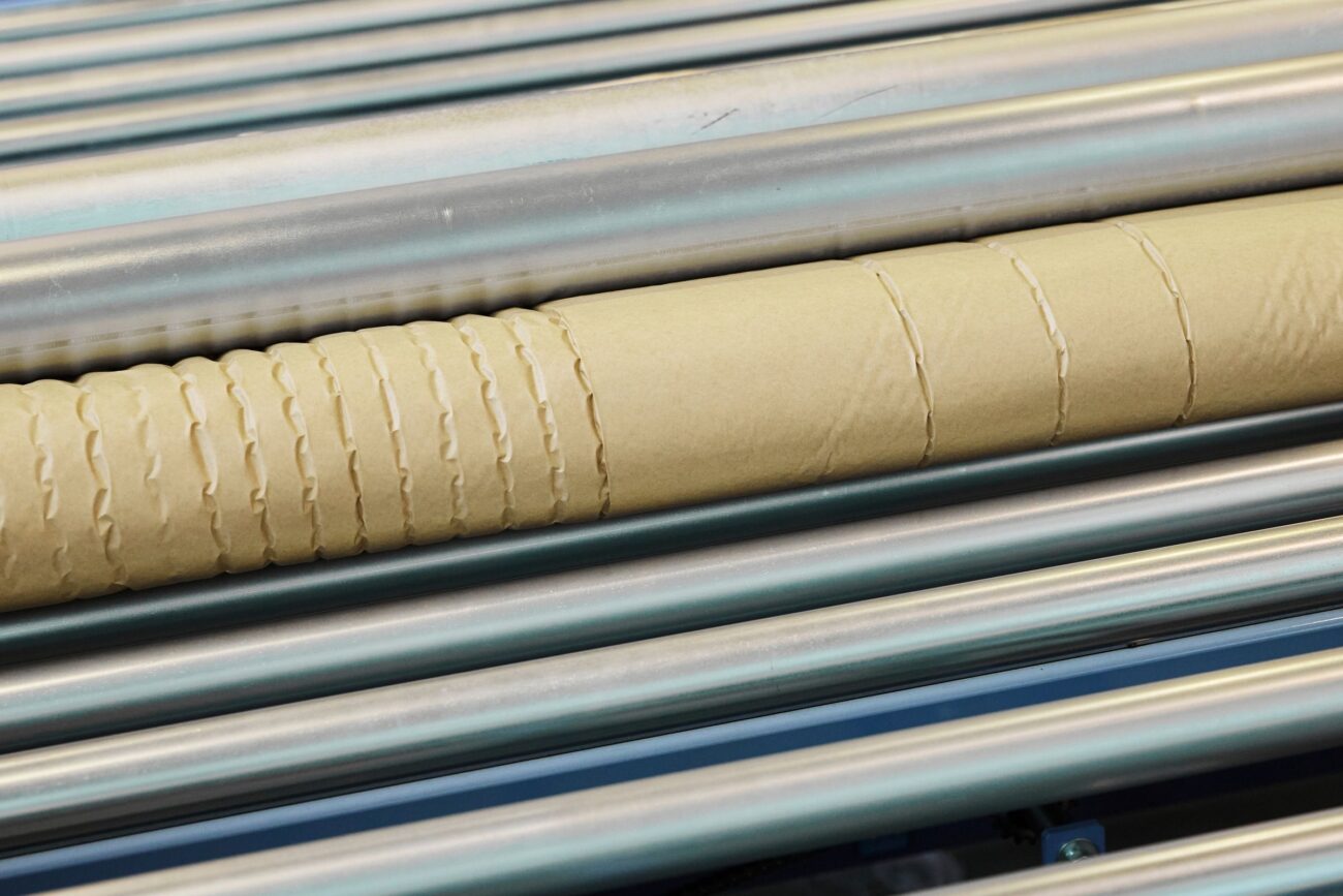 Nahaufnahme einer Papphülse mit gleichmäßig verteilten Rillen, die auf parallelen Metallrollen in einer Produktionsstätte ruht, die sich der Verantwortung und dem Schutz der Umwelt verschrieben hat.