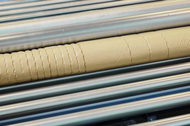 Nahaufnahme einer Papphülse mit gleichmäßig verteilten Rillen, die auf parallelen Metallrollen in einer Produktionsstätte ruht, die sich der Verantwortung und dem Schutz der Umwelt verschrieben hat.