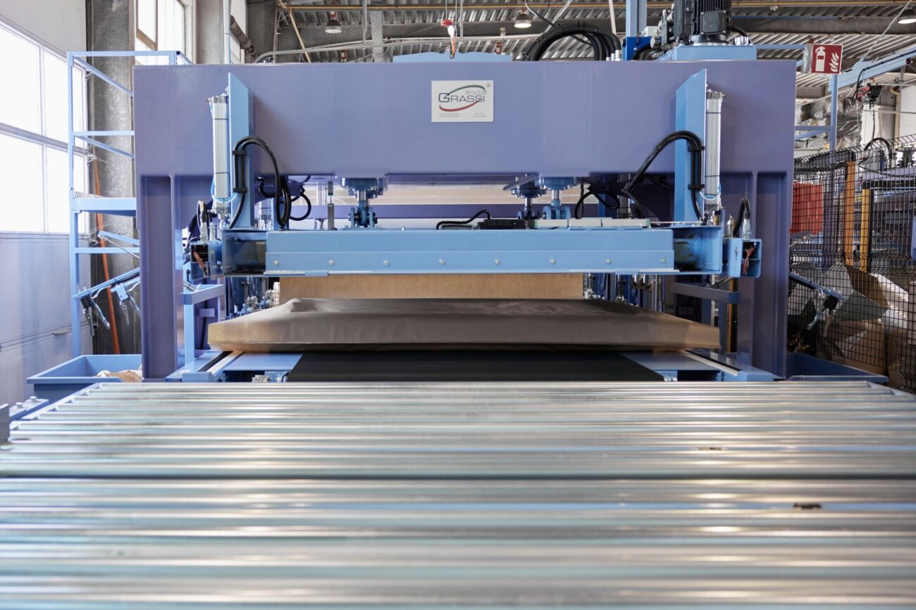 Eine große Industriemaschine mit blauen Komponenten presst Flachmaterial auf ein Förderband in einer Fabrikumgebung, die nachhaltige Verpackung unterstützt. Im Vordergrund sind Metallrollen zu sehen, im Hintergrund weitere Geräte.