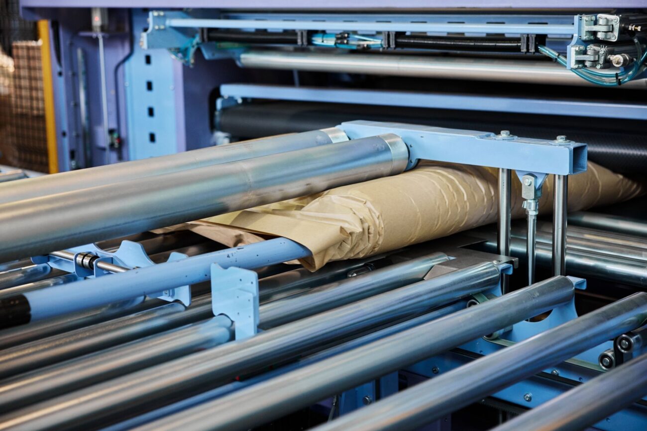 Nahaufnahme einer Verpackungsmaschine, die nachhaltiges braunes Papier um ein Produkt wickelt, wobei Metallrollen und mechanische Teile hervorgehoben werden - ein Beispiel für innovativen Versand und Verpackung mit Schwerpunkt auf Nachhaltigkeit.