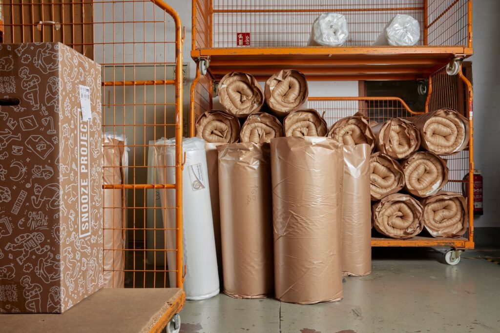 Mehrere aufgerollte und in braunes Papier eingewickelte Matratzen stapeln sich auf orangefarbenen Metallregalen und neben einem Markenkarton und präsentieren nachhaltige Verpackung in einer Lagerhalle mit Betonboden.