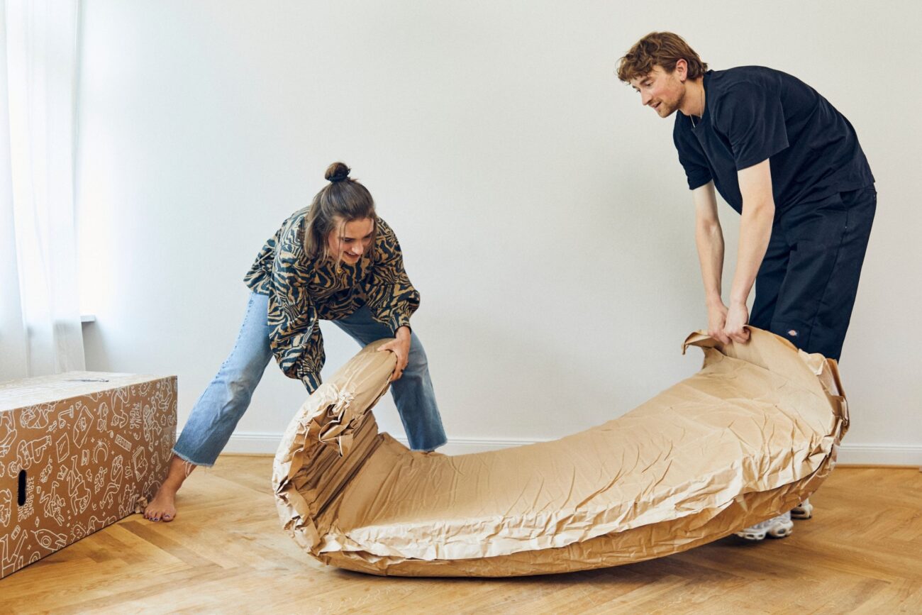 Zwei Personen wickeln gemeinsam eine große, aufgerollte Matratze mit brauner Versandverpackung auf einem Holzboden aus, daneben steht ein offener Karton in einem hellen Raum.