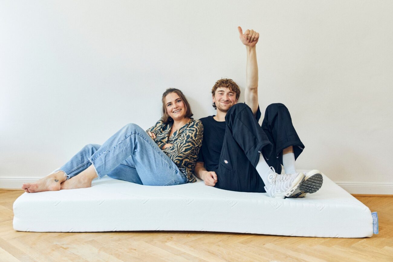 Eine lächelnde Frau und ein Mann sitzen barfuß auf einer Matratze in einem hellen Raum und genießen entspannte Momente. Ihr Glück strahlt, während sie Komfort und Nachhaltigkeit demonstrieren und achtsame Entscheidungen über ihre Umgebung treffen.