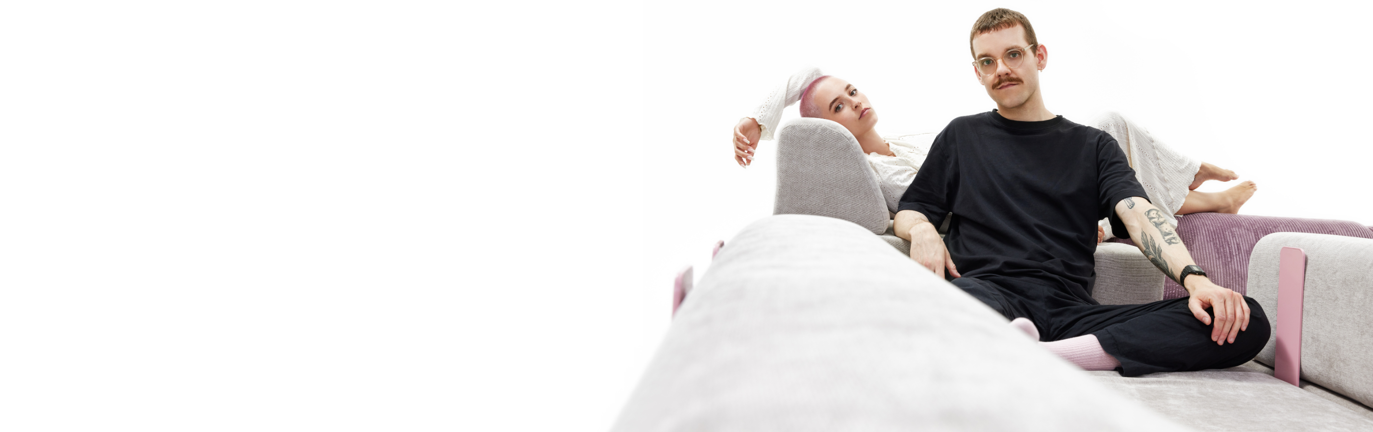 Zwei Personen entspannen sich auf einem modernen, hellen Hero-Sofa; die eine sitzt im Schneidersitz in Schwarz vorne, die andere in Weiß und stützt ihren Kopf auf die Armlehne. Der minimalistische Hintergrund erinnert an eine Startseite mit viel weißem Raum.