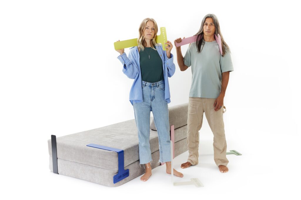 Zwei Personen stehen barfuß neben einem gefalteten Futon und halten jeweils bunte, überdimensionale Messwerkzeuge in der Hand. Mehrere andere Messwerkzeuge liegen neben ihnen auf dem Boden - eine spielerische Annäherung an das Messen für ein Levi Modulares Sofa mit Schlaffunktion. Der Hintergrund ist einfarbig weiß.