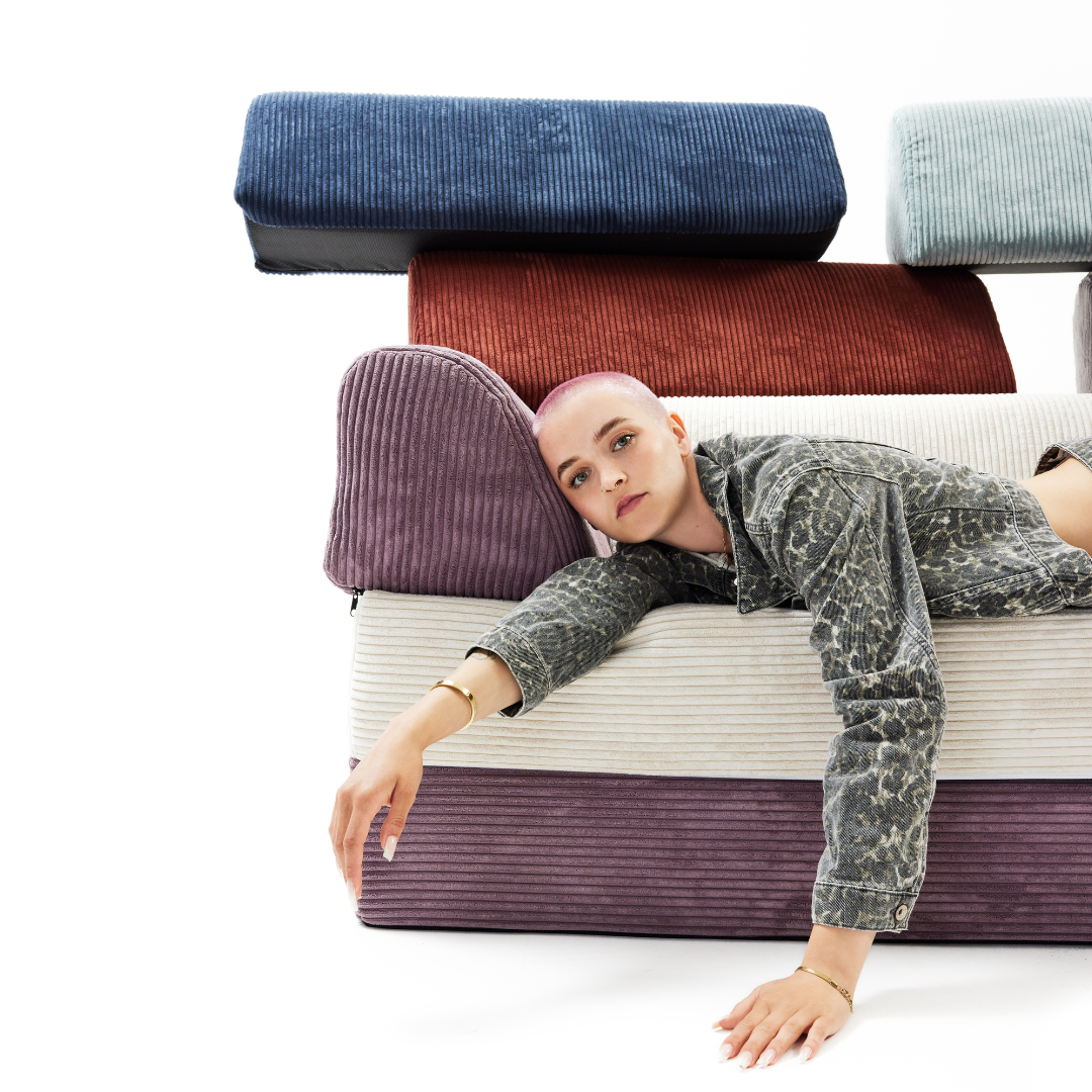 Eine Person mit kurzen Haaren und einer gemusterten "Stoffmuster für Levi"-Jacke lehnt auf gestapelten Cordkissen in Creme, Lavendel, Rost, Blau und Hellblau vor einem schlichten weißen Hintergrund.