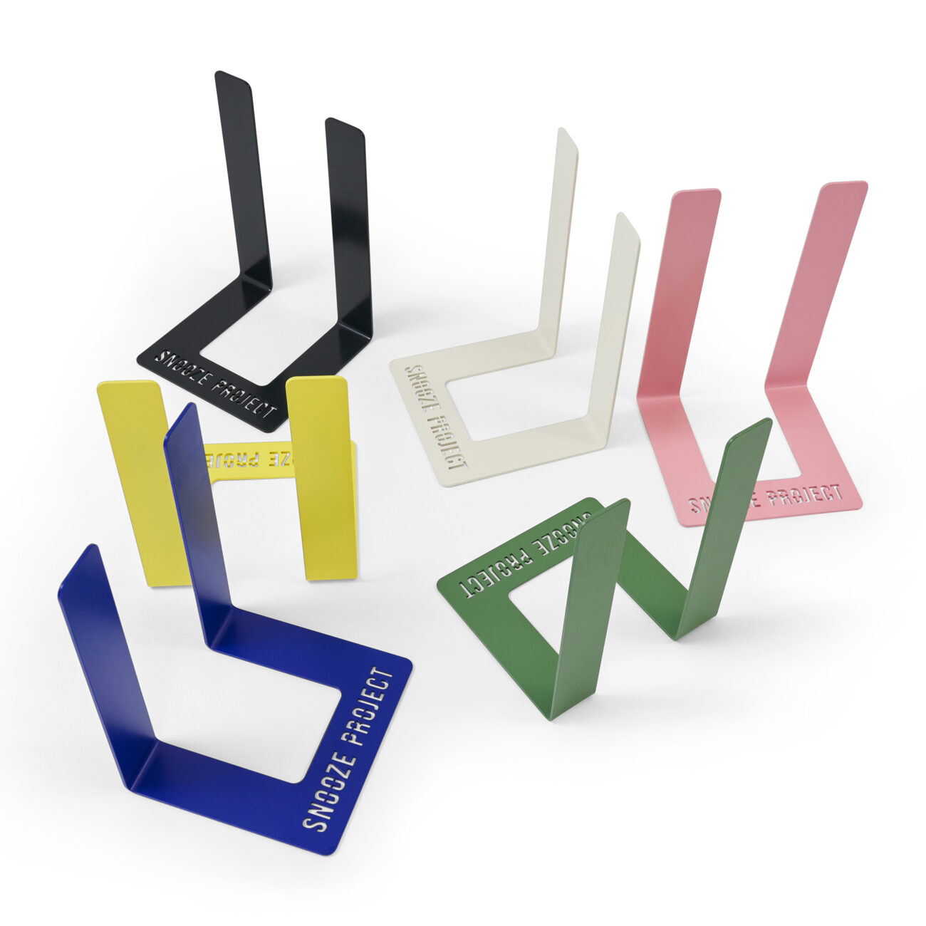 SP_Levi_U-Gestell_01 Fünf metallene Buchstützen in Schwarz, Gelb, Weiß, Rosa, Blau und Grün sind auf einer weißen Fläche angeordnet. Auf jeder Buchstütze ist das SNOOZE PROJECT ausgeschnitten.