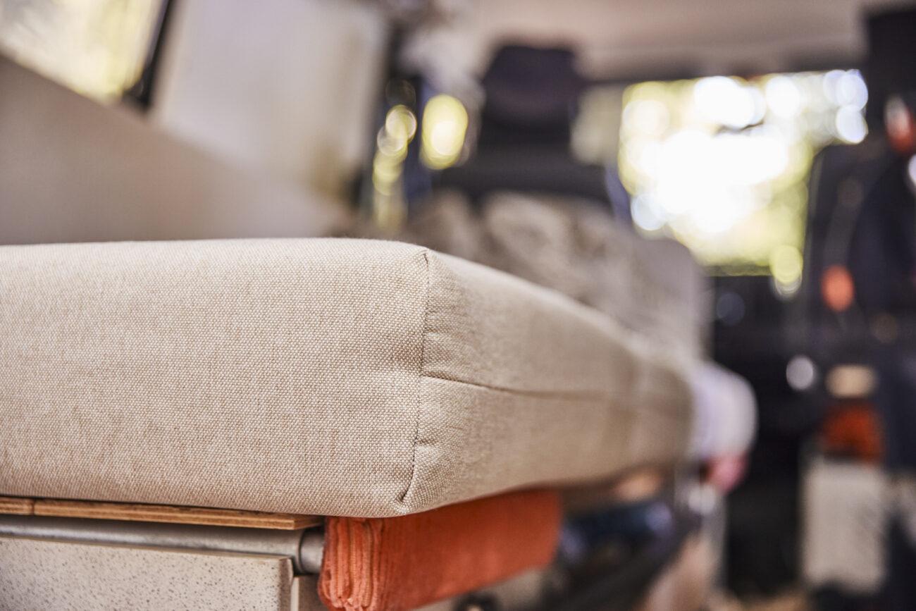 Nahaufnahme eines beigen Polsters nach Maß auf einer Bank in einem Wohnmobil, wobei der Hintergrund leicht verschwommen ist und die Sitze und das durch die Fenster einfallende Sonnenlicht zeigt - eine Einladung zu einer echten Erlebniswelt.