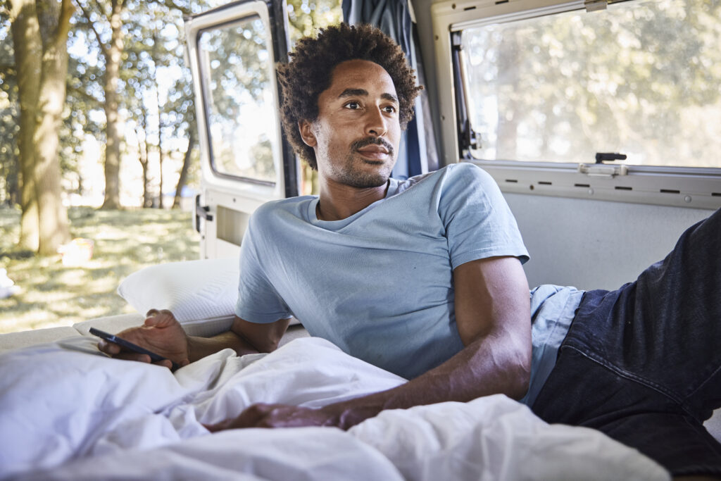 Ein Mann im hellblauen Hemd liegt auf einem Bett mit Polster nach Maß in einem Wohnmobil, hält ein Smartphone in der Hand und schaut nachdenklich aus der offenen Tür auf die sonnige, bewaldete Erlebniswelt draußen.