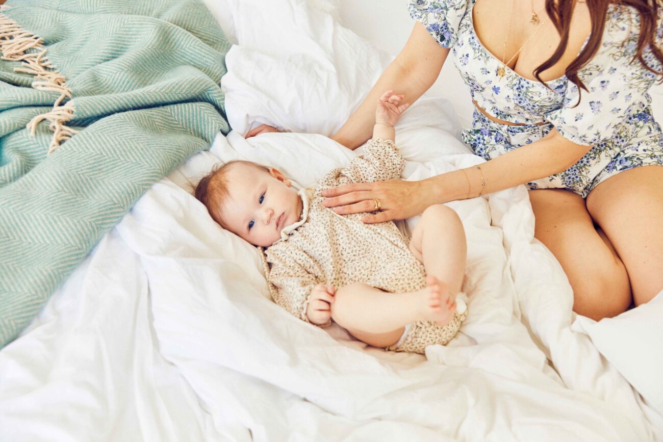 Ein Baby in einem hellen Outfit liegt auf weißen Matratzen in einem Familienbett und schaut auf. Eine erwachsene Frau in einem geblümten Kleid sitzt in der Nähe, ihr Arm berührt sanft das Baby. Eine grüne Decke ist in dieser liebevollen Erlebniswelt zu sehen.