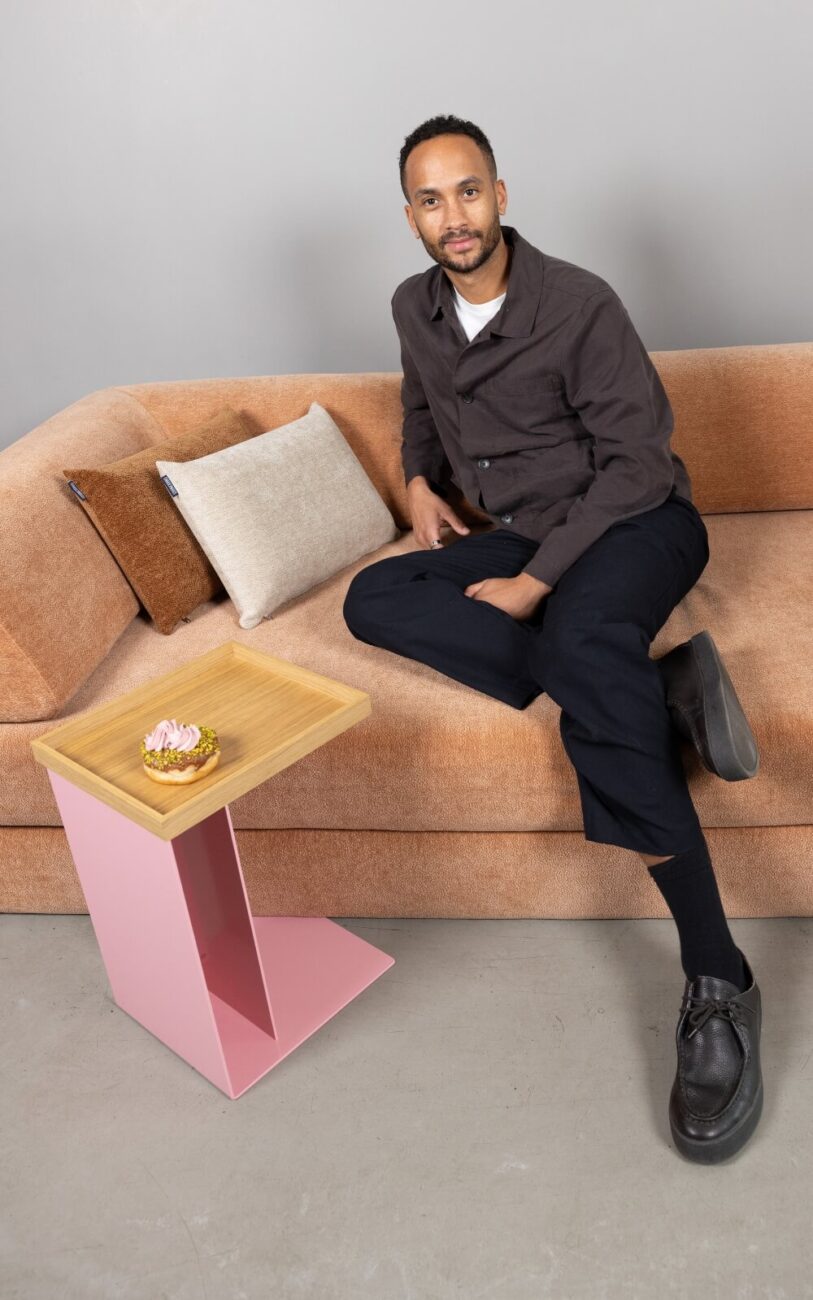Ein Mann sitzt auf einem hellbraunen Sofa Levi mit beigen und braunen Kissen. Er stützt seinen Arm auf die Rückenlehne und schaut in die Kamera. Vor ihm steht ein kleiner rosafarbener Beistelltisch mit einem Gebäck, der vor einem schlichten grauen Hintergrund eine gemütliche Erlebniswelt schafft.
