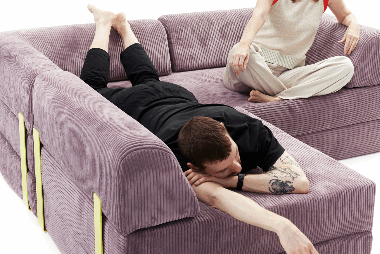 Ein schwarz gekleideter Mann liegt mit dem Gesicht nach unten auf einem lila Cord-Eckschlafsofa und streckt seinen Arm aus. Eine Frau in heller Kleidung sitzt hinter ihm. Das klobige, moderne Sofa dient auch als gemütlicher Schlafplatz - perfekt als Ecksofa-Bett.
