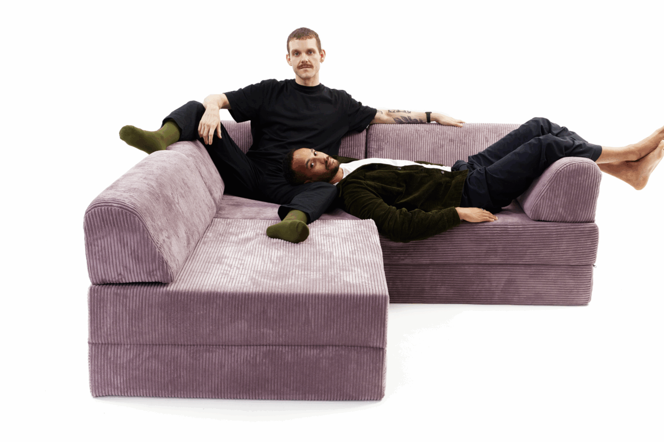 Zwei Personen entspannen sich auf einem hellvioletten Eckschlafsofa. Die eine sitzt aufrecht in schwarzer Kleidung, während die andere mit dem Kopf auf dem Schoß der ersten Person liegt und eine dunkle Hose und ein grünes Oberteil trägt. Der Hintergrund ist einfarbig weiß.