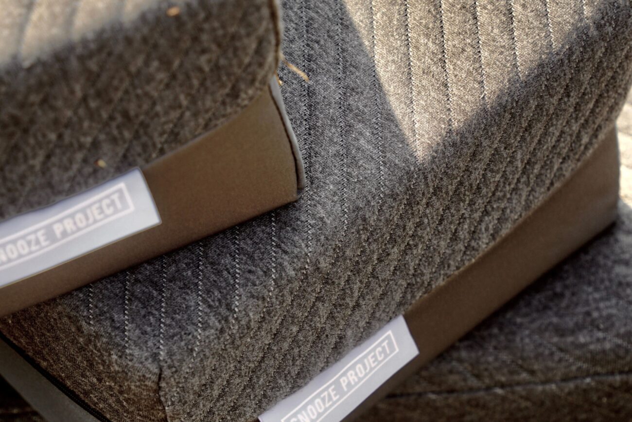 Nahaufnahme von zwei grauen, strukturierten Sitzen mit braunem Rand, die jeweils ein weißes SNOOZE PROJECT Label tragen. Der weiche Stoff hat ein diagonales, fischgrätenartiges Muster - perfekt für jede Matratzen nach Maß Erlebniswelt.