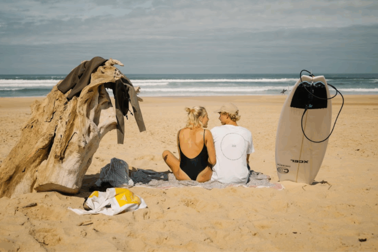 Ein Paar sitzt auf einem Handtuch am Strand mit Blick aufs Meer und genießt seine eigene kleine Erlebniswelt. In der Nähe liegen ein Surfbrett und Treibholz; sie trägt einen schwarzen Badeanzug, er ein weißes Hemd und eine Kappe. Der Himmel ist teilweise bewölkt.