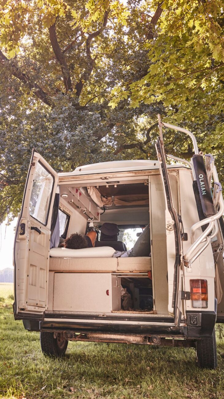 Eine Person liegt in einem Wohnmobil mit geöffneten Hecktüren, das auf einer Wiese unter großen, belaubten Bäumen geparkt ist. Das Sonnenlicht dringt durch die Äste, hebt das individuelle Polster nach Maß hervor und schafft eine friedliche Erlebniswelt im Freien.