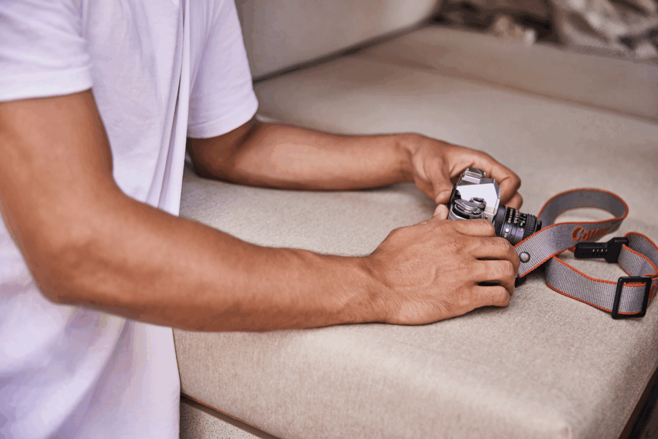 Eine Person in einem weißen T-Shirt hält eine alte Kamera mit einem rot-grauen Kameragurt und stützt ihre Arme auf ein beigefarbenes Polster nach Maß, was die gemütliche Erlebniswelt noch verstärkt.