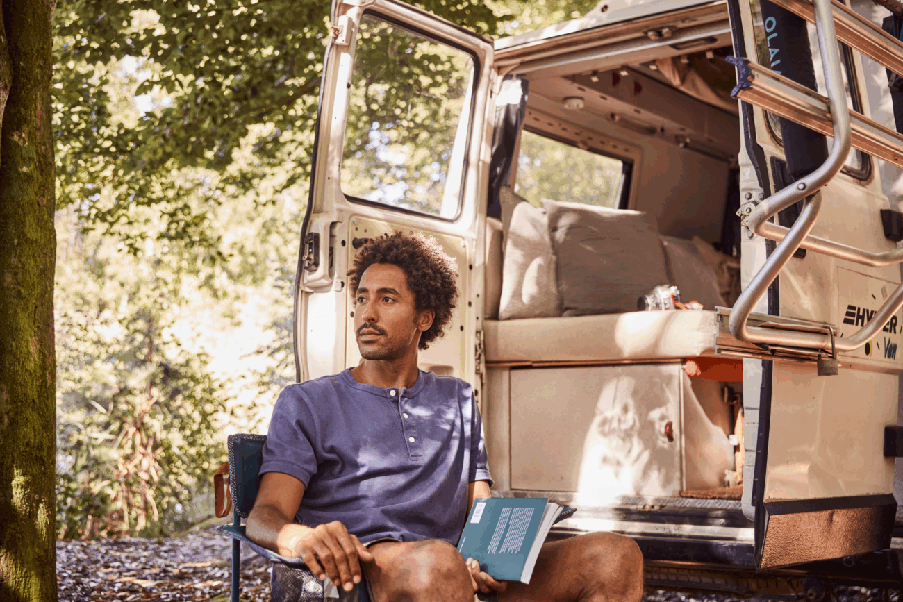 Eine Person sitzt auf einem Stuhl vor einem Wohnmobil in einem Waldgebiet, hält ein Buch in der Hand und schaut nachdenklich zur Seite. Die Tür des Wohnmobils ist offen und zeigt Polster nach Maß und Sonnenlicht, das durch die Bäume fällt.
