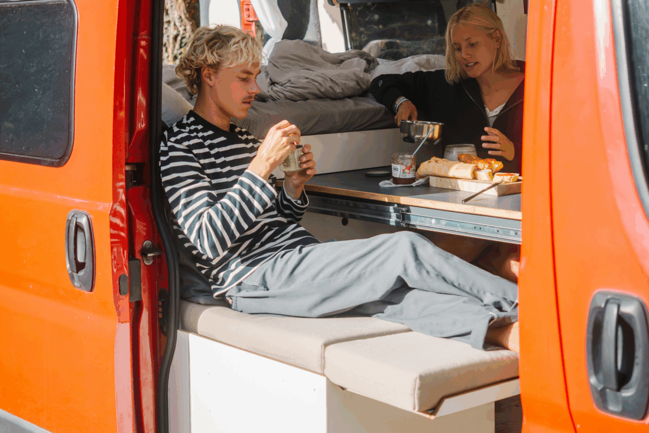 Zwei Personen sitzen in einem roten Wohnmobil mit offener Seitentür und genießen eine Mahlzeit an einem kleinen Tisch. Im Hintergrund ist ein gemütliches Polster nach Maß zu sehen. Der eine schmiert Brot, der andere hält ein Glas und schafft so seine eigene Erlebniswelt auf Rädern.