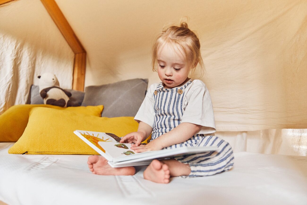 Ein kleines Kind mit blondem Haar in Zöpfen sitzt auf einem Familienbett in einem gemütlichen Zelt und liest ein Bilderbuch. Das Bett hat gelbe Kissen und ein Plüschtier im Hintergrund und schafft eine Erlebniswelt zum Wohlfühlen.
