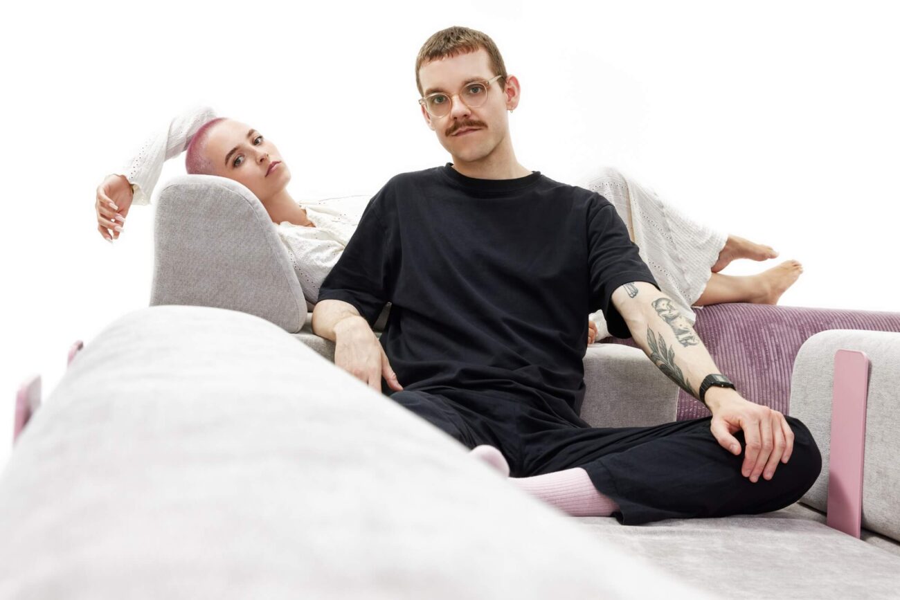 Ein Mann in schwarzer Kleidung und mit Brille sitzt im Schneidersitz auf einem modernen Sofa, während sich eine Frau in einem weißen Kleid hinter ihm zurücklehnt und ihren Kopf auf die Armlehne des Sofas stützt. Der Hintergrund ist weiß und minimalistisch.