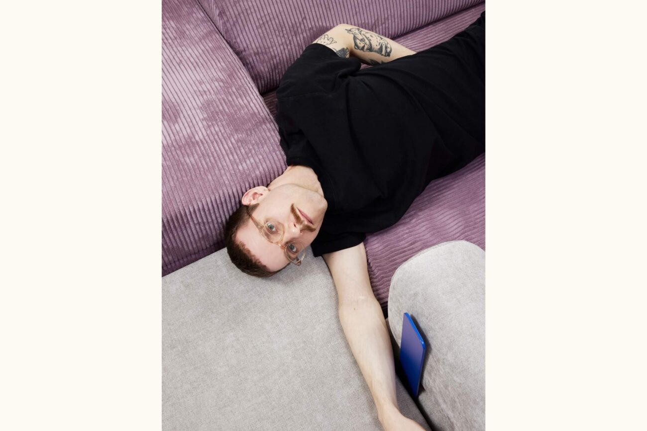 Eine Person mit kurzen Haaren, Brille und Tattoos liegt auf einer lila Kordsofa und trägt ein schwarzes Hemd. Ihr Arm ist ausgestreckt, und ein blaues Telefon liegt zwischen den Polstern.