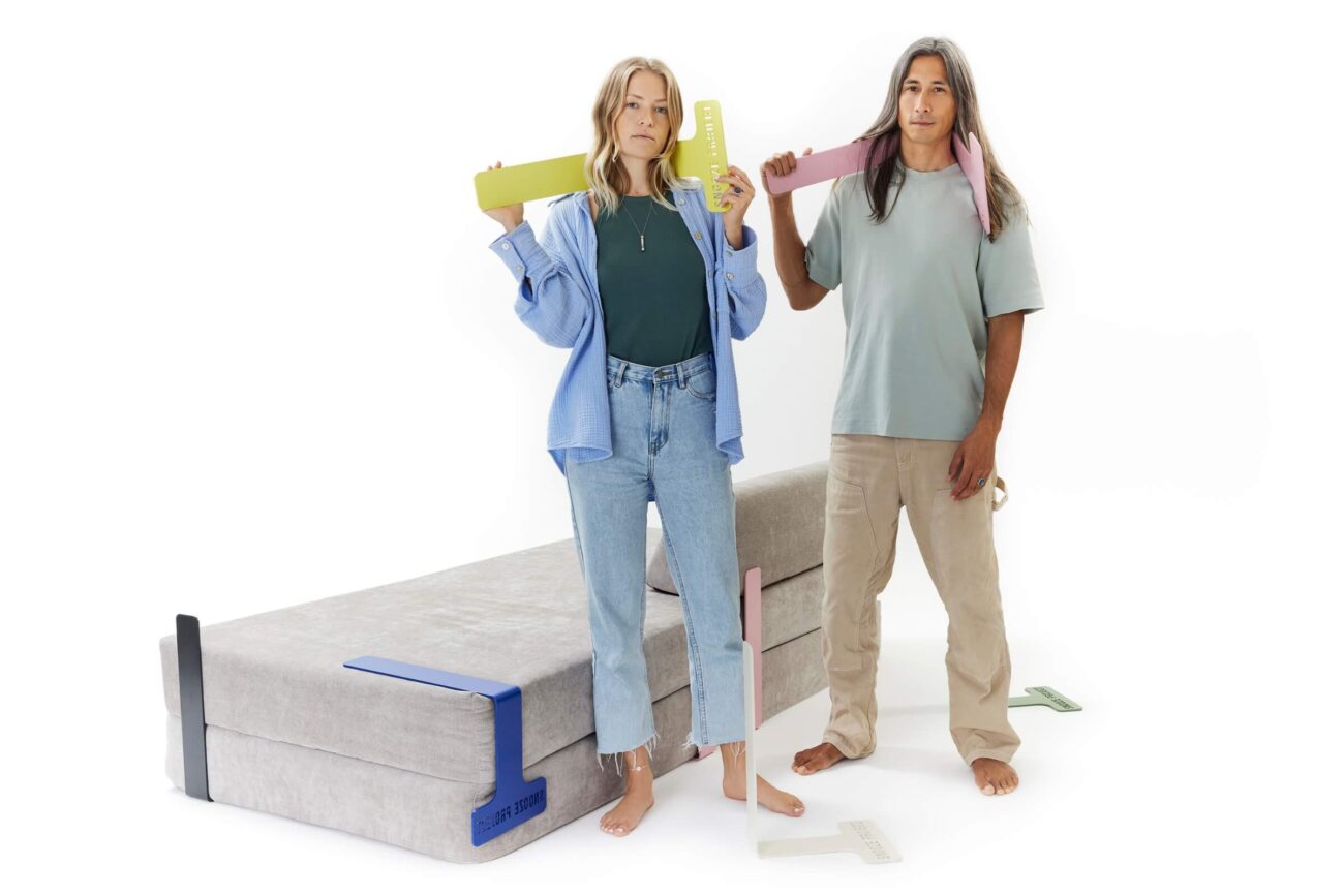 Zwei Personen stehen barfuß neben einem zusammengeklappten grauen Sofa und halten jeweils bunte, große Messwerkzeuge in der Hand. Die Frau trägt Jeans und ein blaues Hemd, der Mann eine beige Hose und ein helles T-Shirt. Messwerkzeuge liegen ebenfalls auf dem Boden.