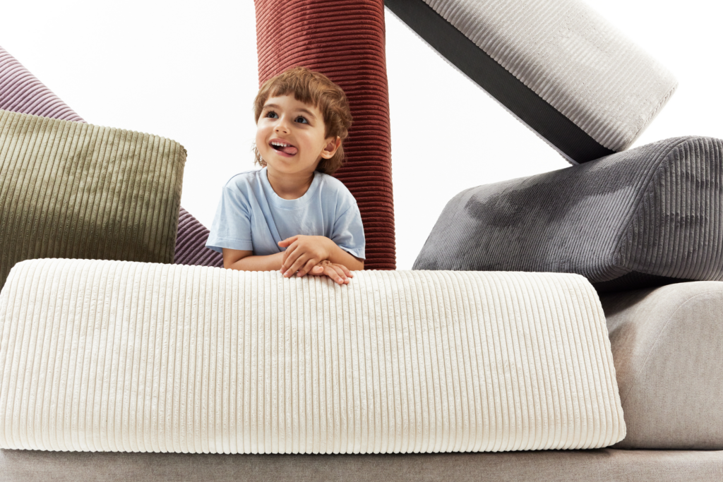 Ein lächelndes kleines Kind in einem hellblauen Hemd ist umgeben von großen, bunten, kordartigen Kissen, die in verschiedenen Formen und Richtungen angeordnet sind - eine ideale Inspiration für jede Schlafsofa-Kategorieseite. Der Hintergrund ist hell und weiß.