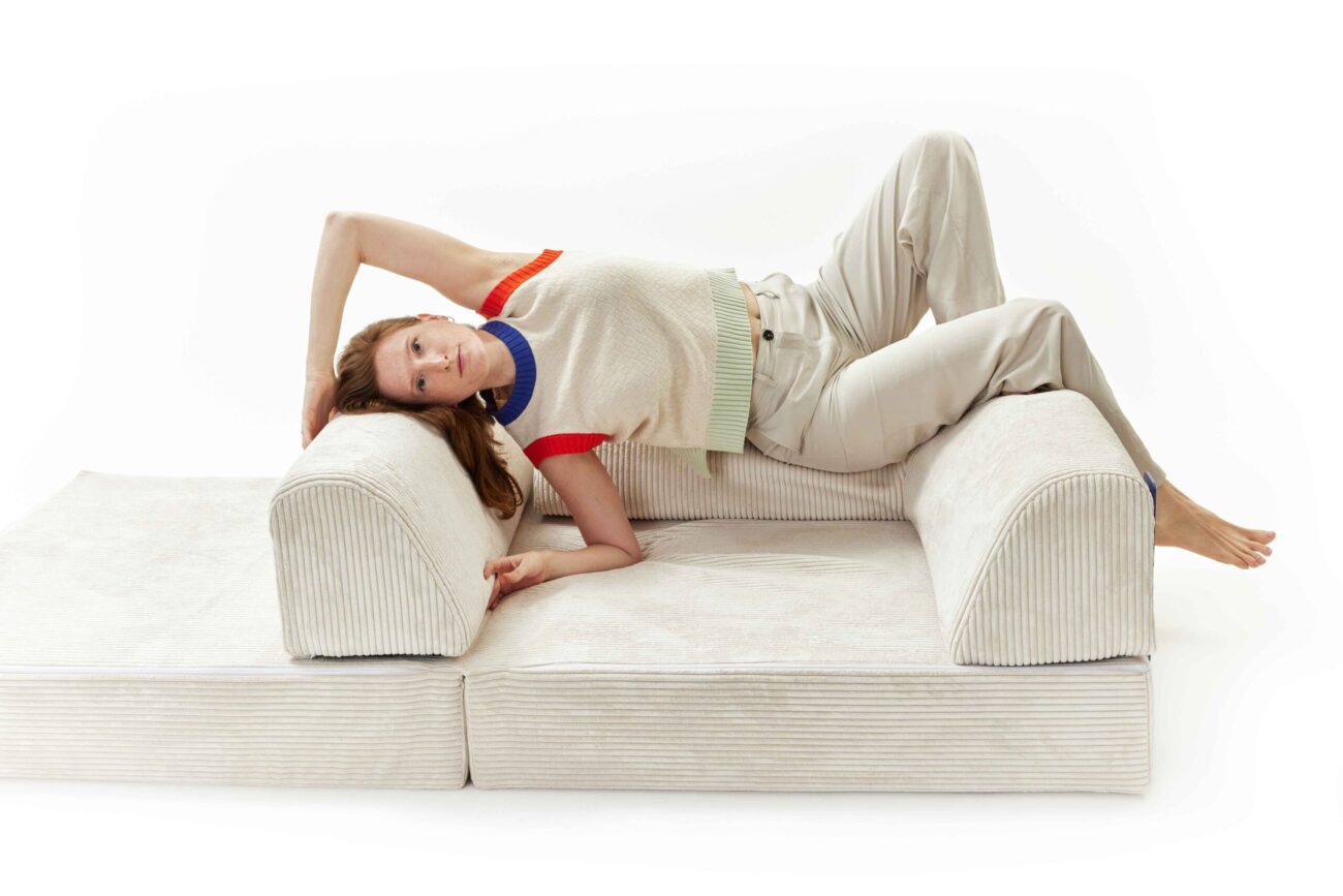 Eine Frau in heller Hose und ärmellosem Pullover liegt seitlich auf dem modularen Sofa Levi, den Kopf auf eine Armlehne gestützt, die Beine über die andere gebeugt. So entsteht eine heitere Erlebniswelt vor einem strahlend weißen Hintergrund.
