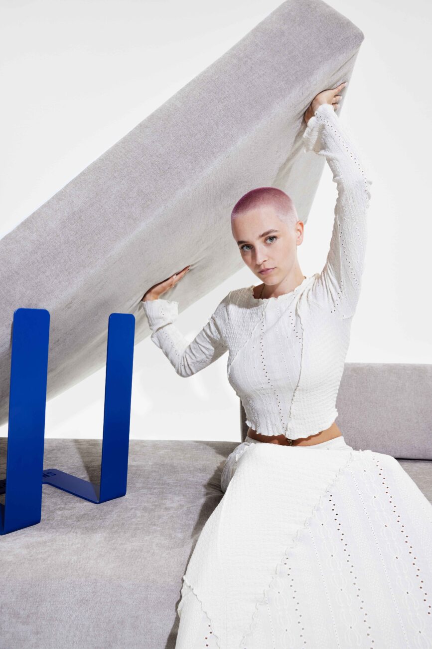 Eine Frau mit kahlgeschorenem Kopf in einem langen weißen Kleid hebt ein großes, hellgraues Sofa Levi-Kissen, das von einem blauen Metallrahmen getragen wird, vor einem schlichten weißen Hintergrund an - und erweckt damit die Erlebniswelt des modernen Designs zum Leben.