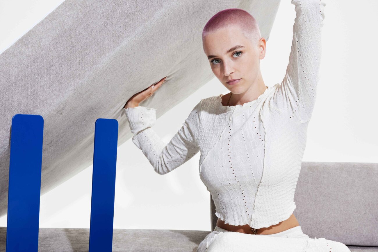 Eine Person mit rasiertem, pastellrosa Kopf und hellen Augen, die ein strukturiertes weißes Outfit trägt, posiert in einem Haus und hält ein großes, gekipptes Sofa-Levi-Kissen mit blauen rechteckigen Objekten davor hoch.