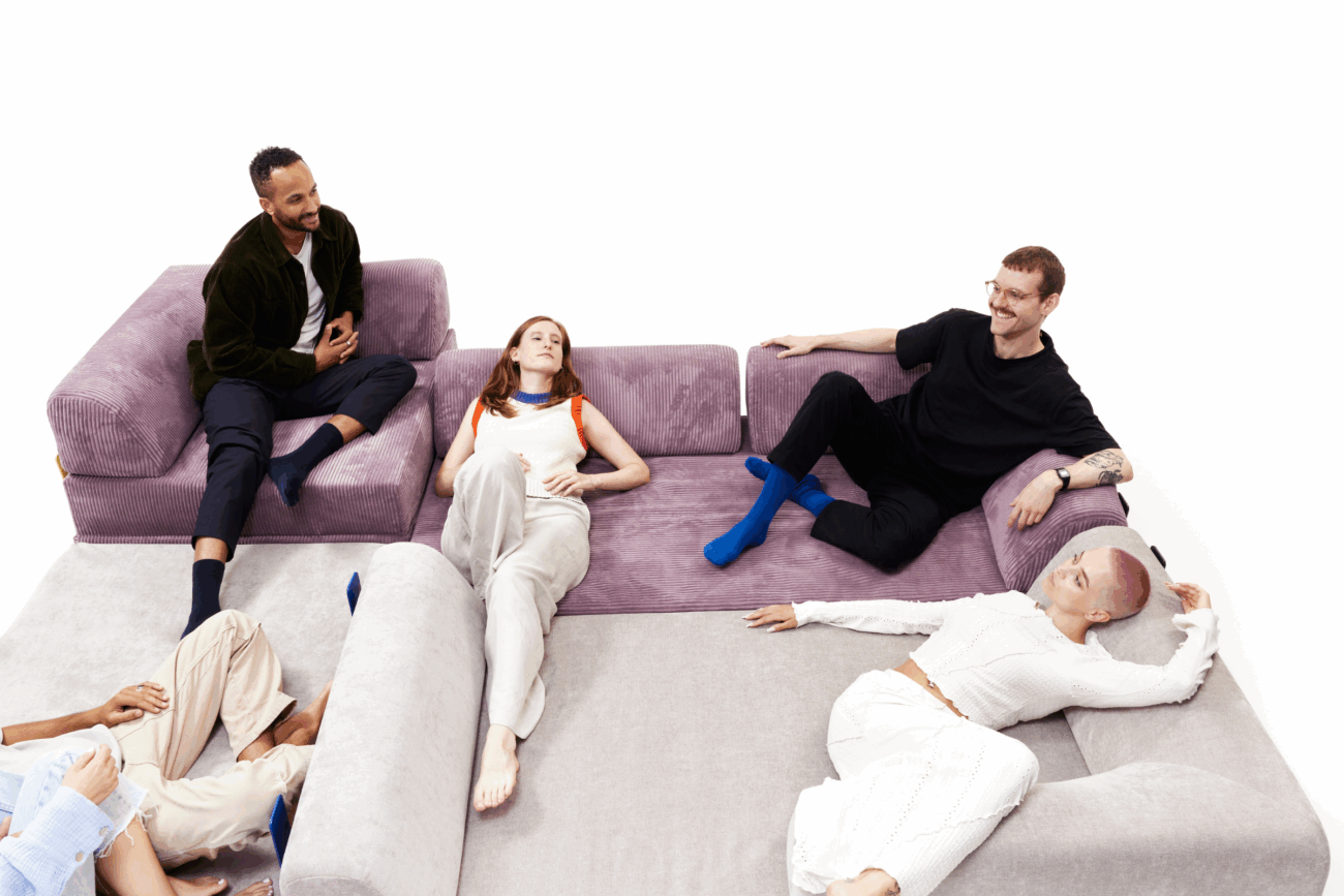 Vier Personen entspannen sich auf einem großen, hellvioletten Schlafsofa in einem weißen Raum, wobei zwei aufrecht sitzen und zwei liegen und sich zu unterhalten und die Gesellschaft des anderen zu genießen scheinen. Perfekt für jede Kategorieseite, die auf Komfort und Stil setzt.