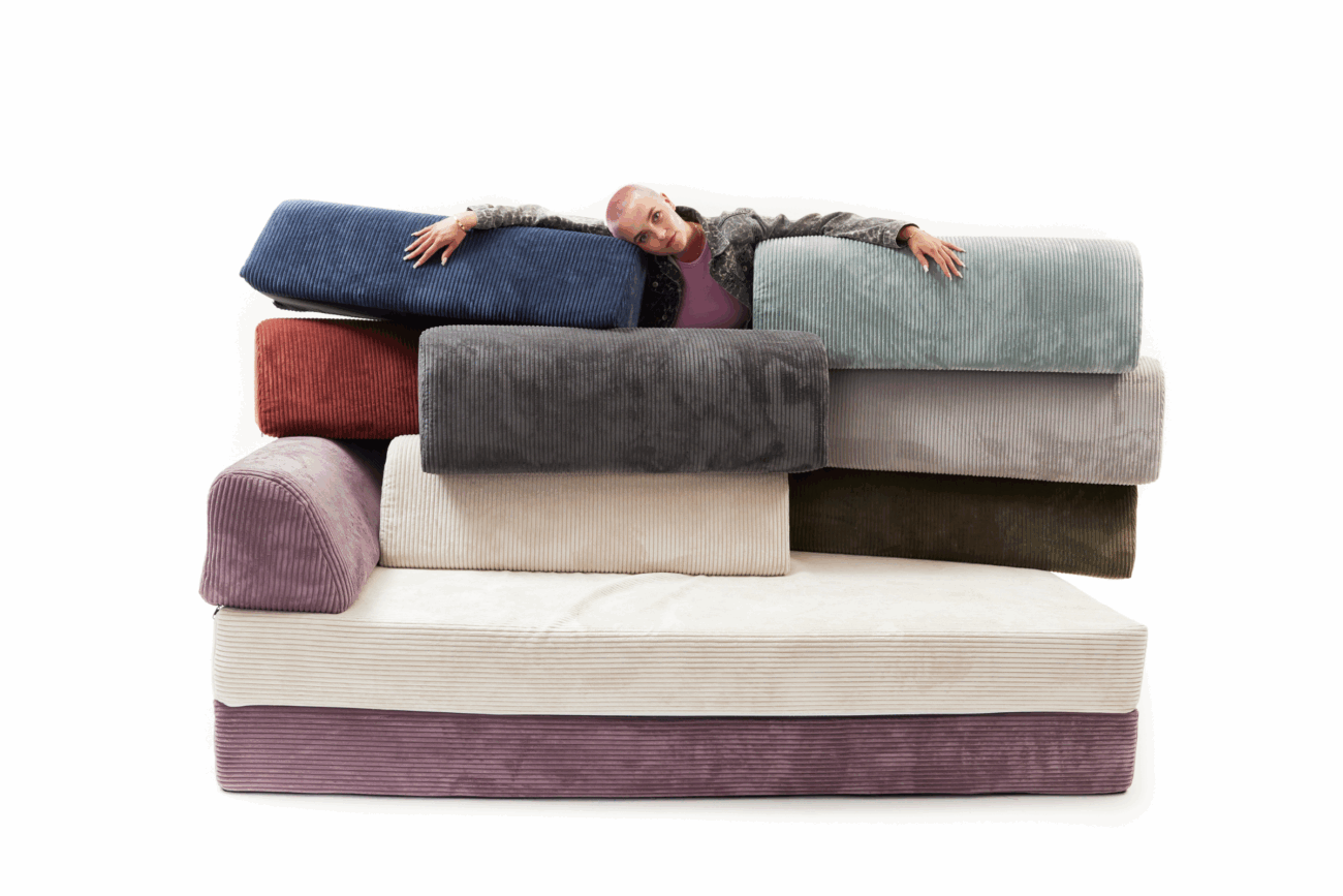 Eine Person mit rasiertem Kopf lächelt, während sie sich über einen Stapel bunter Cord-Schlafsofakissen beugt, die in einem ungleichmäßigen Stapel vor einem weißen Hintergrund angeordnet sind, perfekt für jede Kategorieseite.