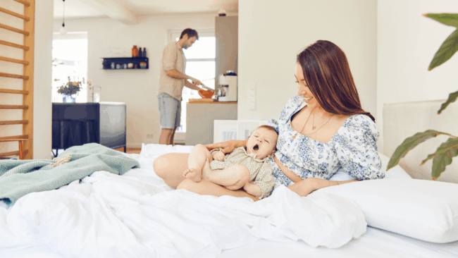 Eine Frau sitzt auf einem Bett und lächelt ein gähnendes Baby auf ihrem Schoß an, während ein Mann in der Küche ihres hellen, gemütlichen Hauses Essen zubereitet und einen friedlichen Morgen im März 2025 genießt.