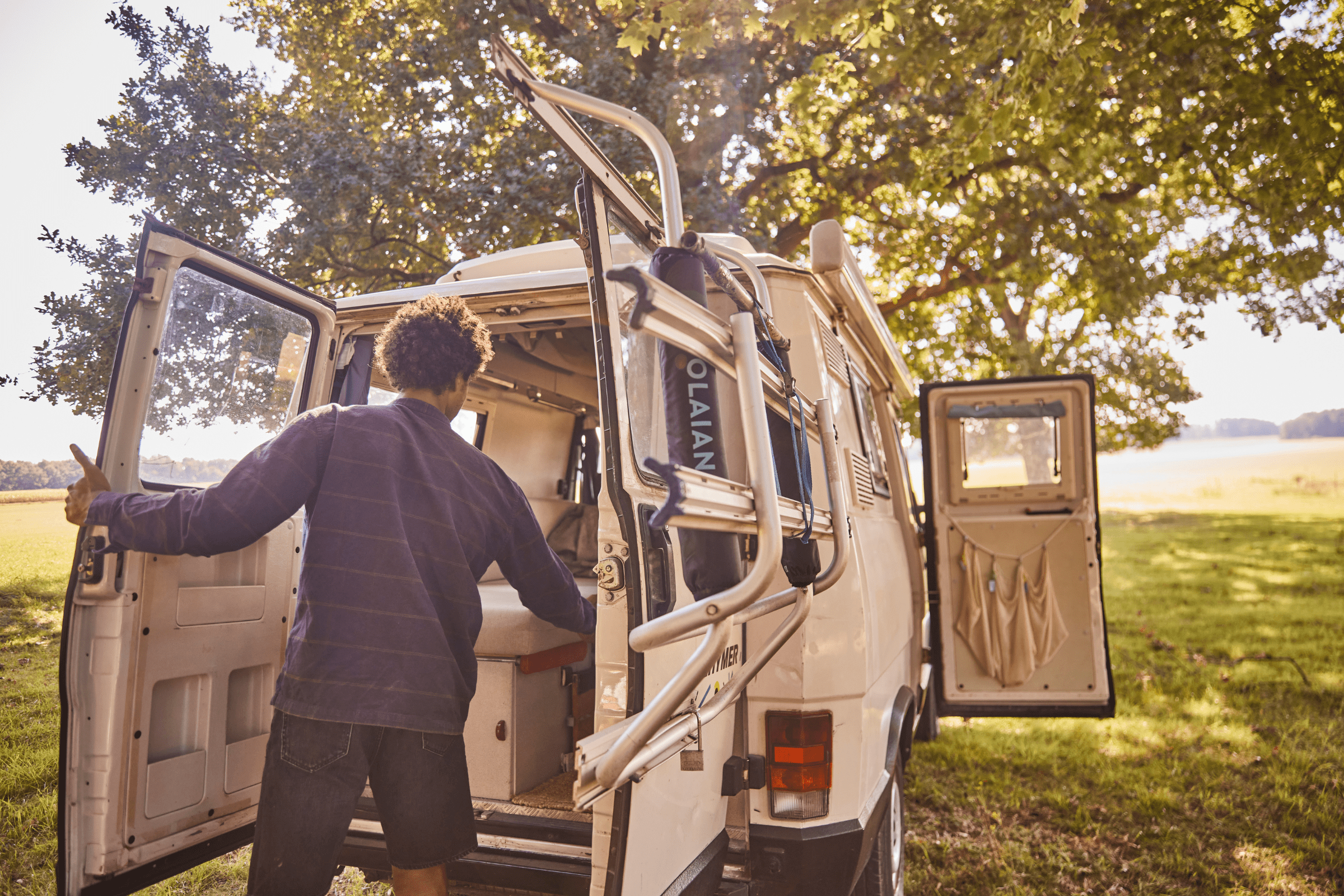Eine Person steht an der offenen Hintertür eines weißen Wohnmobils, das unter einem großen Baum geparkt ist. Das Sonnenlicht dringt durch die Blätter und ein grasbewachsenes Feld erstreckt sich in der Ferne.