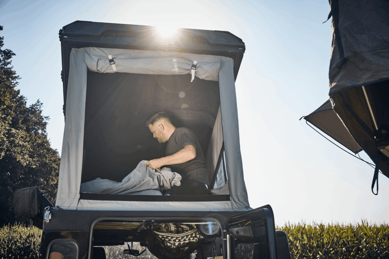 Ein Mann richtet das Bettzeug in einem auf einem Fahrzeug montierten Dachzelt her, während über ihm das Sonnenlicht scheint und im Hintergrund Grünflächen zu sehen sind.