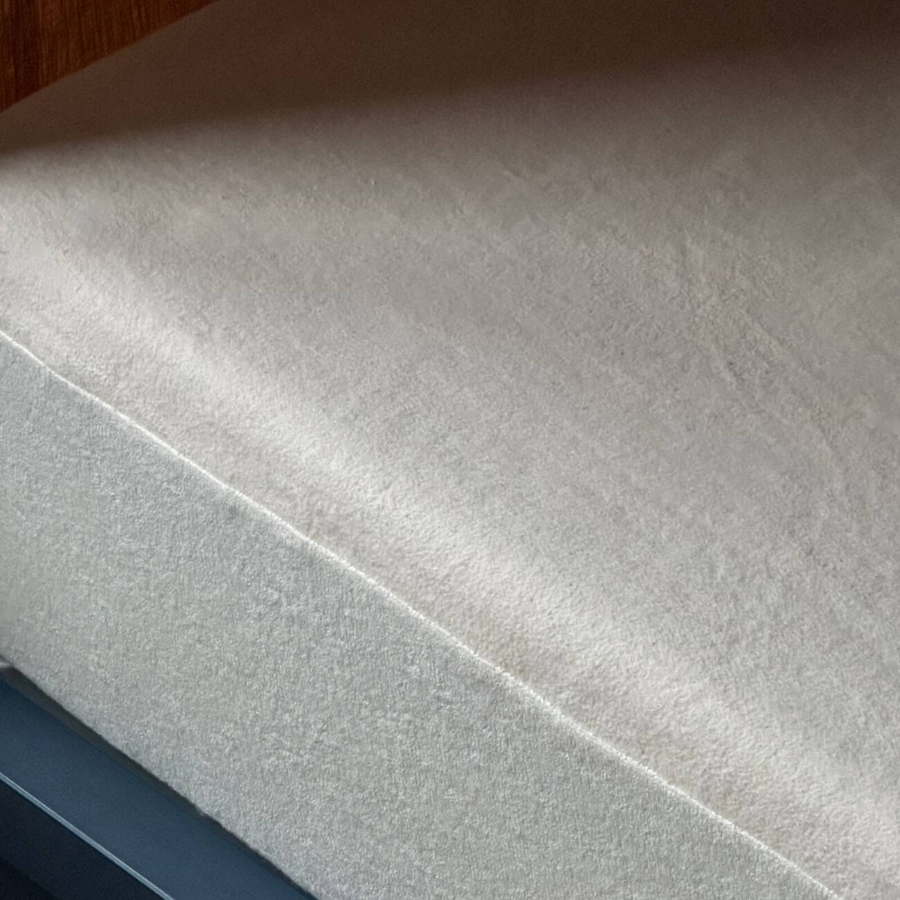 Erlebniswelt_Matratzenschoner-nach-Maß_KONF_04 Eine Nahaufnahme eines ordentlich gemachten Bettes mit einem einfarbigen, hellen Spannbettlaken und einem Matratzenschoner nach Maß, der die Matratze bedeckt. Das Bettgestell ist dunkel, und ein Teil einer Holzwand trägt zur Erlebniswelt im Hintergrund bei.